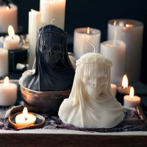 May include: Two white and black skull-shaped candles with a crown on top, surrounded by other candles on a wooden tray.