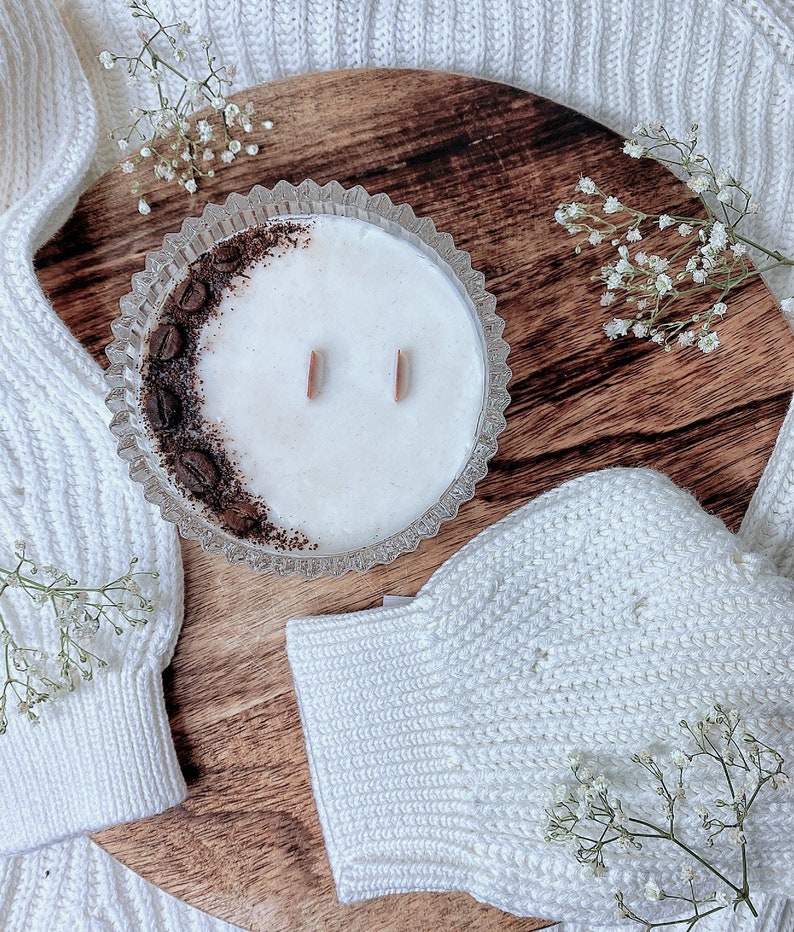 May include: A white candle with a coffee bean design on the side, in a clear glass jar with a decorative rim. The candle is sitting on a wooden surface with white knitted fabric.