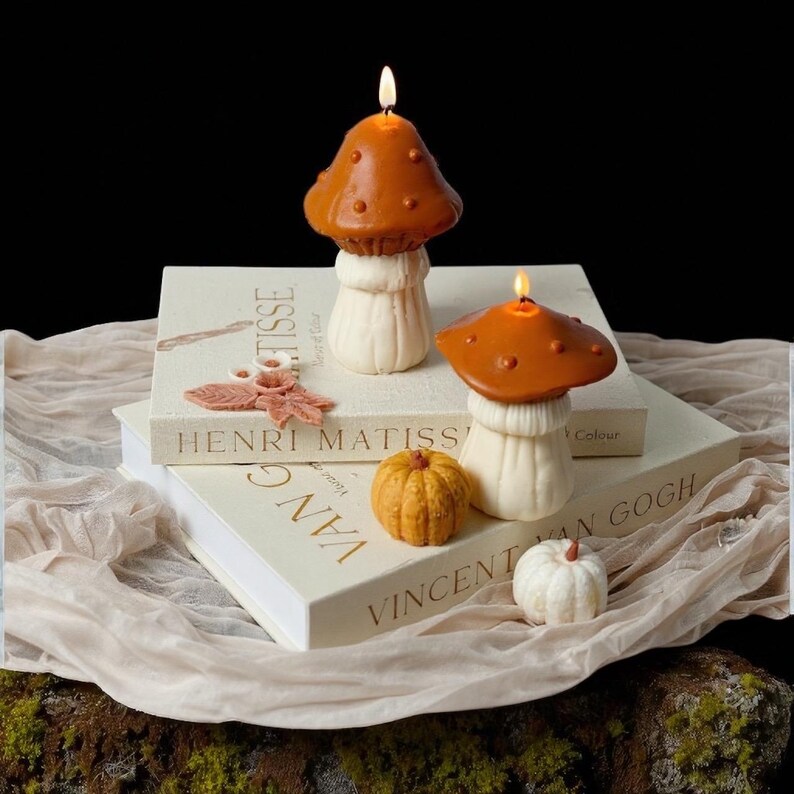 May include: Two lit mushroom-shaped candles with brown caps and white stems sit atop books with the titles "Henri Matisse" and "Vincent Van Gogh." A small orange pumpkin and a white pumpkin are also present. The background is dark.