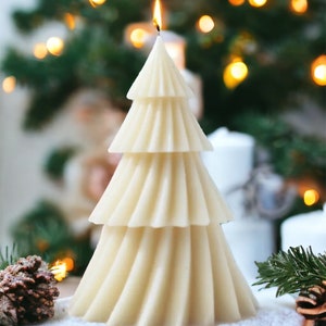 May include: A white, three-tiered, tree-shaped candle with a lit wick, sitting on a wooden log with snow-like white powder around it. The candle is in front of a blurred background of a Christmas tree with lights.
