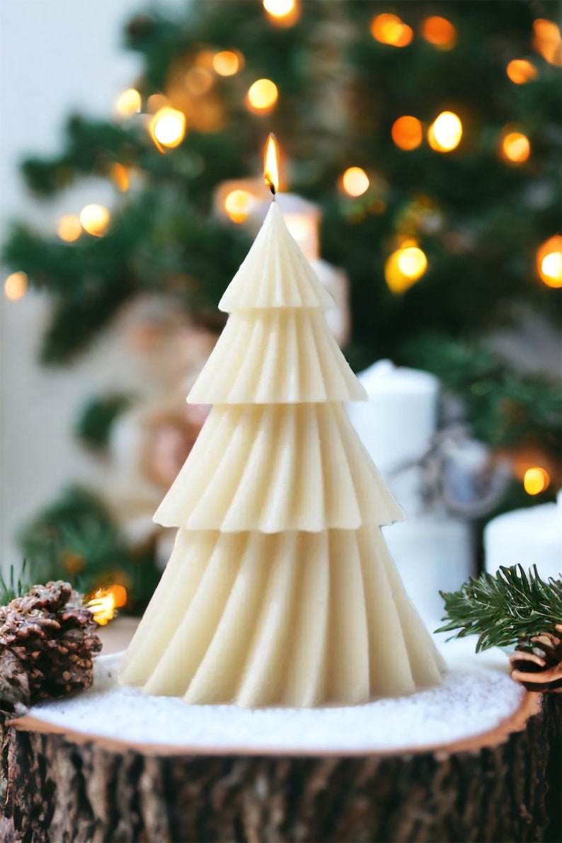 May include: A white, three-tiered, tree-shaped candle with a lit wick, sitting on a wooden log with snow-like white powder around it. The candle is in front of a blurred background of a Christmas tree with lights.