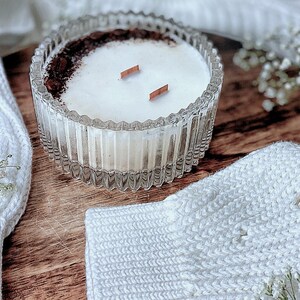 May include: A white candle with a brown rim and two wooden wicks in a clear glass jar with a textured design. The candle is sitting on a wooden surface with white knitted fabric and white flowers.