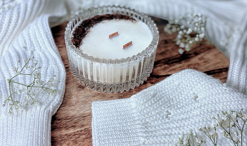 May include: A white candle with a brown rim and two wooden wicks in a clear glass jar with a textured design. The candle is sitting on a wooden surface with white knitted fabric and white flowers.