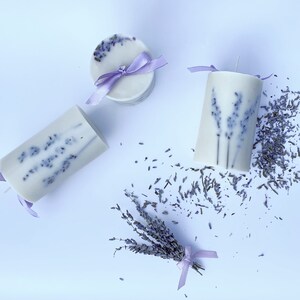 May include: Three lavender scented candles with dried lavender flowers. Two candles are cylindrical and one is round. All candles have a purple ribbon tied around them.