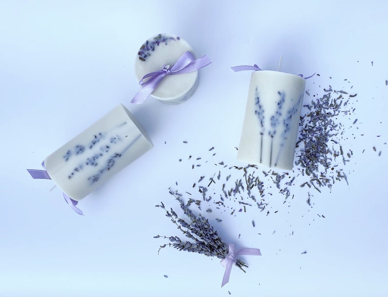 May include: Three lavender scented candles with dried lavender flowers. Two candles are cylindrical and one is round. All candles have a purple ribbon tied around them.