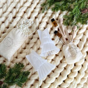 May include: Two clear plastic Christmas tree candle molds, two small brown glass bottles, two wooden honey dippers, and two white muslin bags filled with white wax flakes. The items are arranged on a white knitted blanket with green pine sprigs.