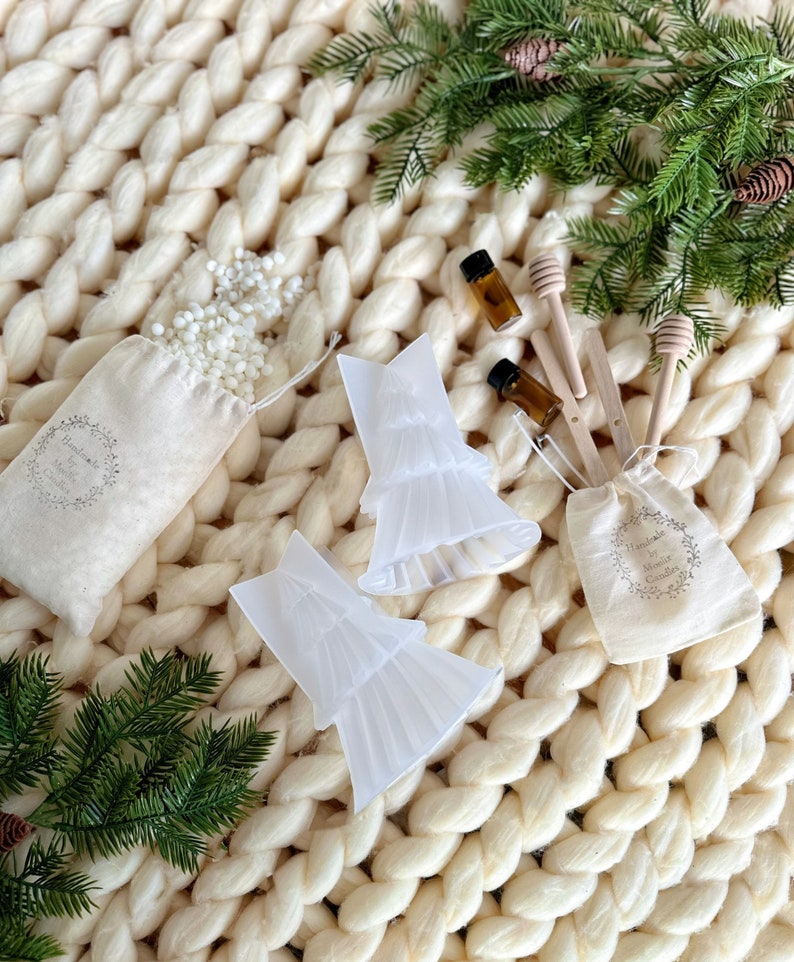May include: Two clear plastic Christmas tree candle molds, two small brown glass bottles, two wooden honey dippers, and two white muslin bags filled with white wax flakes. The items are arranged on a white knitted blanket with green pine sprigs.