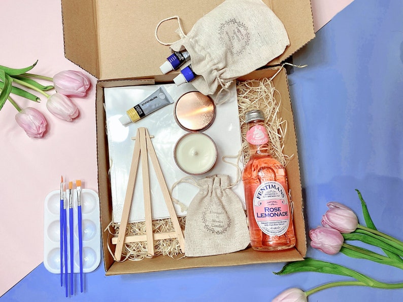 May include: A gift box filled with art supplies, including a small easel, paintbrushes, paint tubes, and a canvas. The box also includes a candle, a bottle of rose lemonade, and a small pouch.