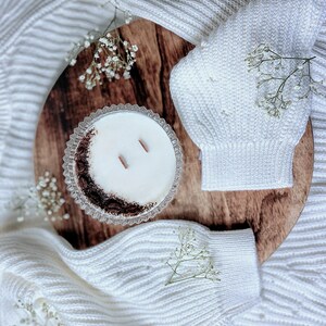 May include: A white candle in a clear glass jar with a brown rim sits on a wooden surface. The candle has two wicks and is surrounded by white flowers.