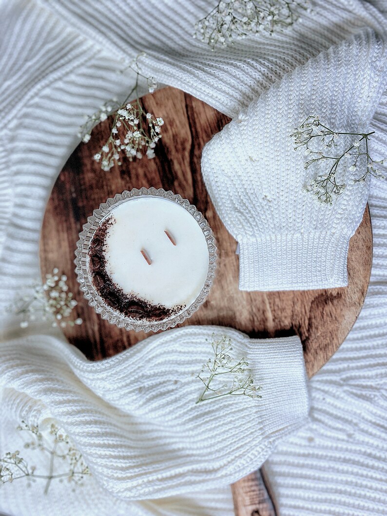 May include: A white candle in a clear glass jar with a brown rim sits on a wooden surface. The candle has two wicks and is surrounded by white flowers.