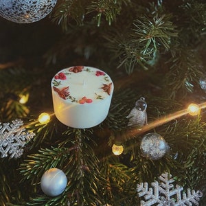 May include: A white candle with dried herbs and red berries on top, sitting on a Christmas tree with silver ornaments and lights.