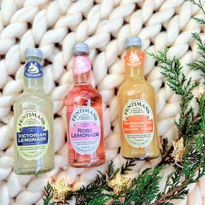 May include: Three glass bottles of Fentimans Botanically Brewed beverages. The bottles are labeled "Victorian Lemonade", "Rose Lemonade", and "Mandarin and Seville Orange Jigger". The bottles are sitting on a white knitted surface with green foliage and gold stars.