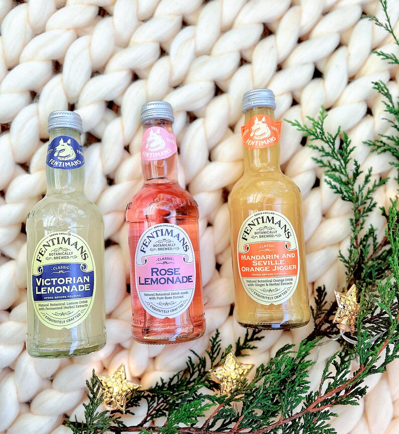 May include: Three glass bottles of Fentimans Botanically Brewed beverages. The bottles are labeled "Victorian Lemonade", "Rose Lemonade", and "Mandarin and Seville Orange Jigger". The bottles are sitting on a white knitted surface with green foliage and gold stars.