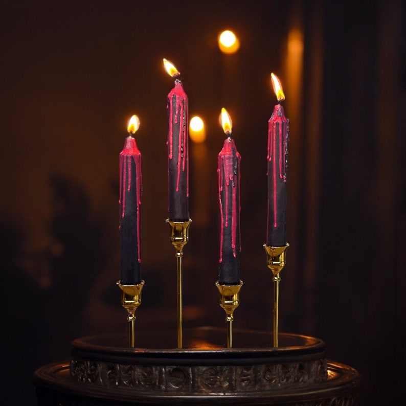 May include: Four black candles with red wax dripping down the sides. The candles are on gold candle holders and are arranged in a row on a dark wooden surface.