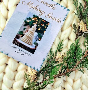 May include: A white booklet with the title "Candle Making Guide" and a photo of a white candle shaped like a Christmas tree. The booklet is on a white fluffy surface with a green branch and gold star ornaments.