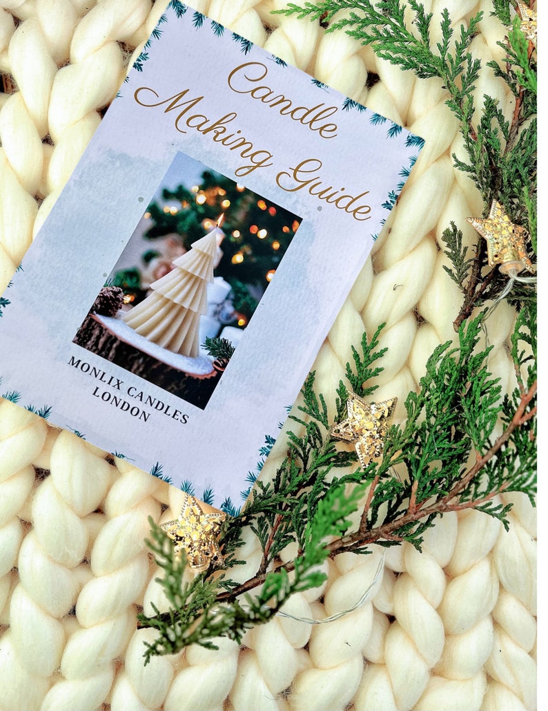 May include: A white booklet with the title "Candle Making Guide" and a photo of a white candle shaped like a Christmas tree. The booklet is on a white fluffy surface with a green branch and gold star ornaments.