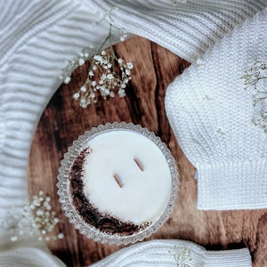 May include: A white candle with a brown coffee bean rim in a clear glass jar with two wooden wicks. The candle is sitting on a wooden surface with a white knitted blanket in the background.