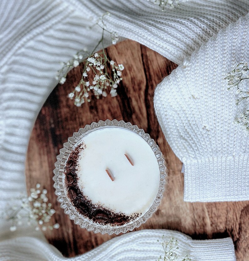 May include: A white candle with a brown coffee bean rim in a clear glass jar with two wooden wicks. The candle is sitting on a wooden surface with a white knitted blanket in the background.