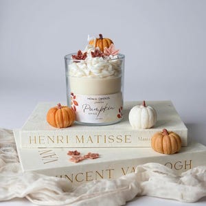 Op de afbeelding: Een glazen kaars met cr&egrave;me-kleurig was en een slagroom-achtige topping, versierd met miniatuur pompoenen en bladeren. Het label luidt "Pumpkin Spice". De kaars staat op een stapel boeken met de namen "Henri Matisse" en "Vincent Van Gogh".