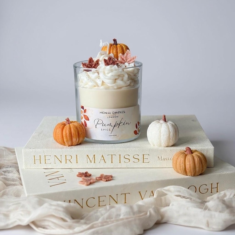 Op de afbeelding: Een glazen kaars met cr&egrave;me-kleurig was en een slagroom-achtige topping, versierd met miniatuur pompoenen en bladeren. Het label luidt "Pumpkin Spice". De kaars staat op een stapel boeken met de namen "Henri Matisse" en "Vincent Van Gogh".