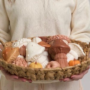 May include: A woven basket filled with mushroom and pumpkin-shaped candles in shades of brown, orange, and white. The basket has two handles and is held by a person wearing a cream-colored sweater.