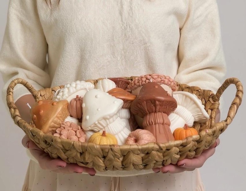 May include: A woven basket filled with mushroom and pumpkin-shaped candles in shades of brown, orange, and white. The basket has two handles and is held by a person wearing a cream-colored sweater.