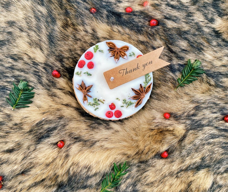 May include: A white candle with dried flowers and star anise on a brown faux fur background. The candle has a brown wooden base and a small brown tag with the text "Thank you" attached.