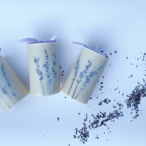 May include: Three white pillar candles with lavender pressed into the wax. The candles are tied with purple ribbon and are on a white background. Dried lavender flowers are scattered around the candles.
