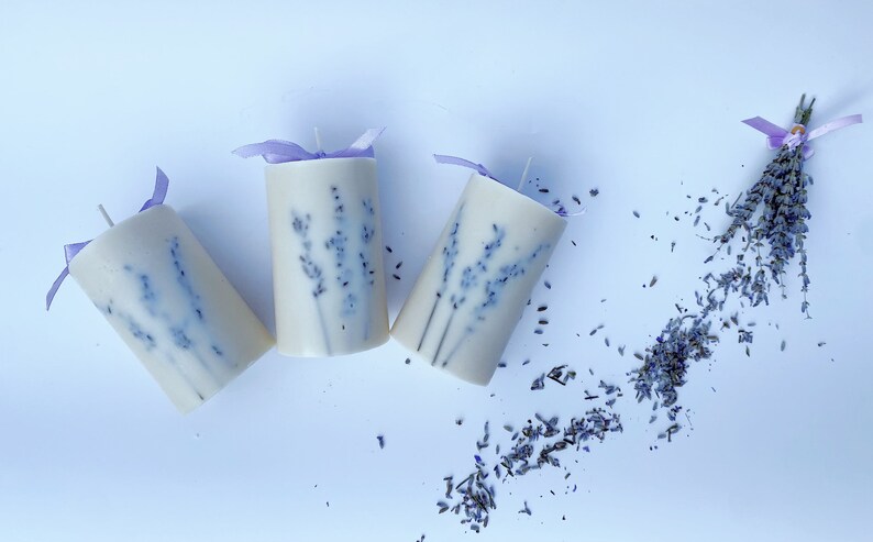 May include: Three white pillar candles with lavender pressed into the wax. The candles are tied with purple ribbon and are on a white background. Dried lavender flowers are scattered around the candles.
