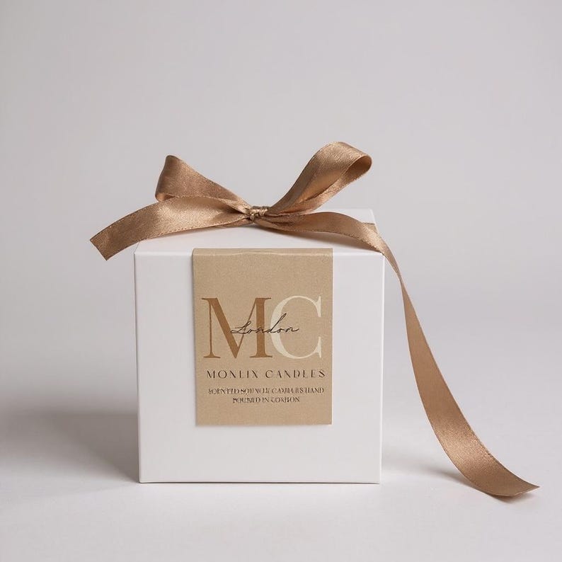 May include: A white square gift box tied with a gold satin ribbon. The box has a brown label with the text "MC London" and "MONLIX CANDLES" printed on it. The ribbon is tied in a bow.