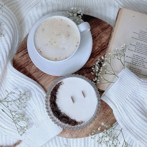 May include: A white ceramic mug filled with a light brown coffee drink with foam on top, a white saucer, a white candle in a clear glass jar with a brown rim, a wooden cutting board, and white flowers on a white knitted blanket. The text "TREES" is visible on a book in the background.