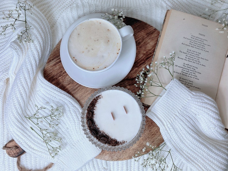 May include: A white ceramic mug filled with a light brown coffee drink with foam on top, a white saucer, a white candle in a clear glass jar with a brown rim, a wooden cutting board, and white flowers on a white knitted blanket. The text "TREES" is visible on a book in the background.