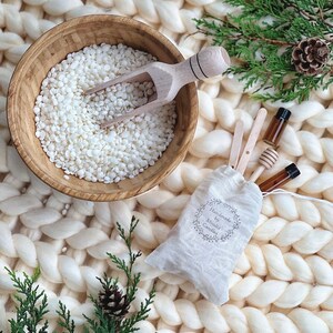 May include: A white bowl filled with white wax flakes and a wooden scoop. A small white bag with the text "Handmade by Monikx Candles" is next to the bowl. Two small glass bottles with brown liquid and wooden sticks are in the bag. The items are on a white fluffy surface.