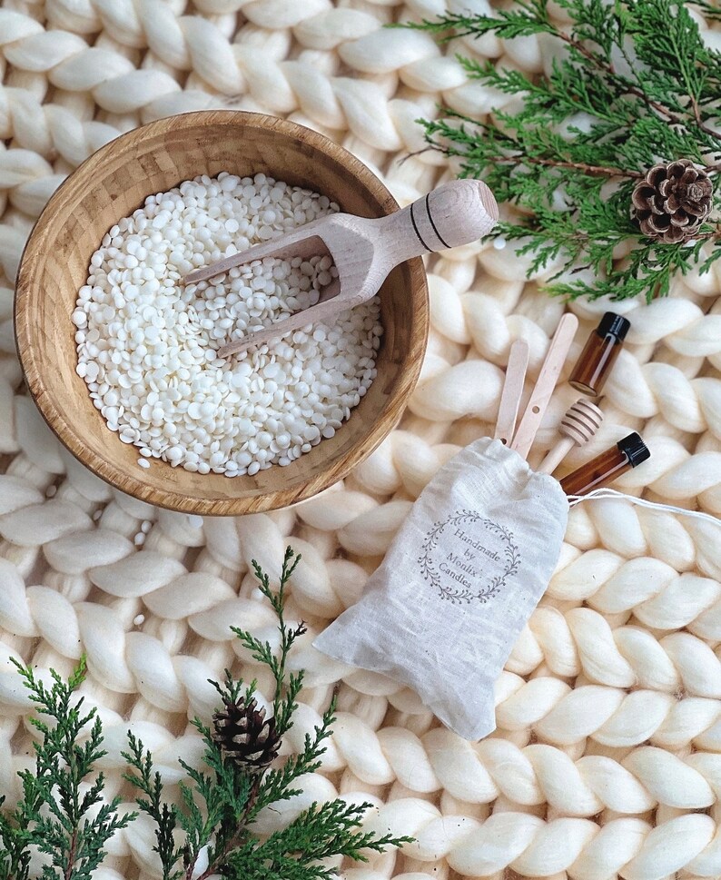 May include: A white bowl filled with white wax flakes and a wooden scoop. A small white bag with the text "Handmade by Monikx Candles" is next to the bowl. Two small glass bottles with brown liquid and wooden sticks are in the bag. The items are on a white fluffy surface.