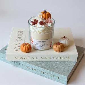 Op de afbeelding: Een gelaagde pompoen kruidkaars in een heldere glazen pot, gegarneerd met slagroomwas, miniatuurpompoenen en herfstbladeren. De kaars staat op een boek getiteld "Vincent Van Gogh". Extra kleine pompoenen zijn rond de kaars geplaatst.