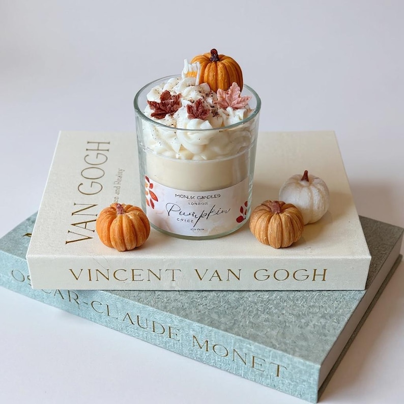 Op de afbeelding: Een gelaagde pompoen kruidkaars in een heldere glazen pot, gegarneerd met slagroomwas, miniatuurpompoenen en herfstbladeren. De kaars staat op een boek getiteld "Vincent Van Gogh". Extra kleine pompoenen zijn rond de kaars geplaatst.