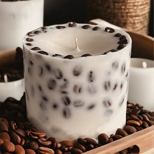 Pu&ograve; includere: Una candela bianca con chicchi di caff&egrave; incorporati nella cera. La candela &egrave; appoggiata su un vassoio di legno con chicchi di caff&egrave; sparsi intorno.