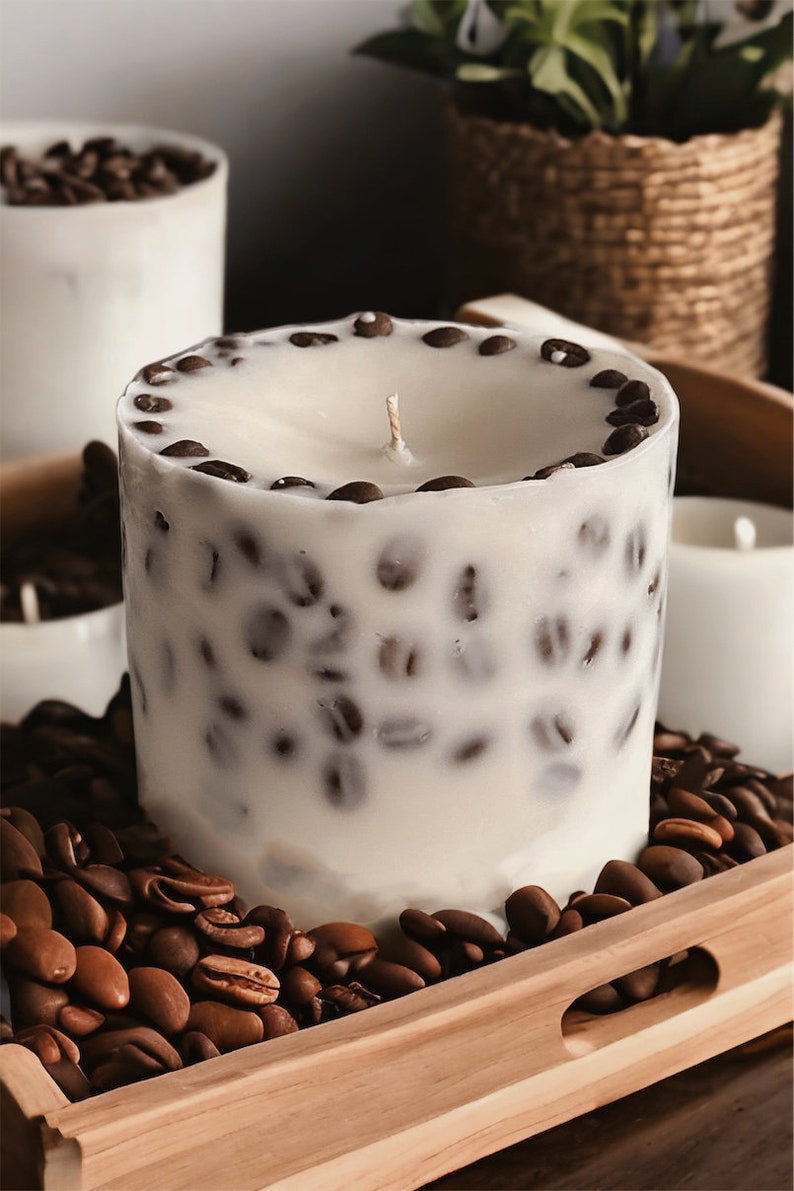Pu&ograve; includere: Una candela bianca con chicchi di caff&egrave; incorporati nella cera. La candela &egrave; appoggiata su un vassoio di legno con chicchi di caff&egrave; sparsi intorno.