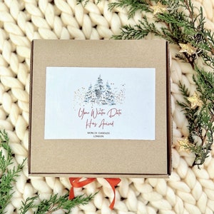 May include: A brown cardboard box with a white label that says "Your Winter Date Has Arrived" and "MONIX CANDLES LONDON". The box is sitting on a white knitted blanket with green pine branches and gold stars.
