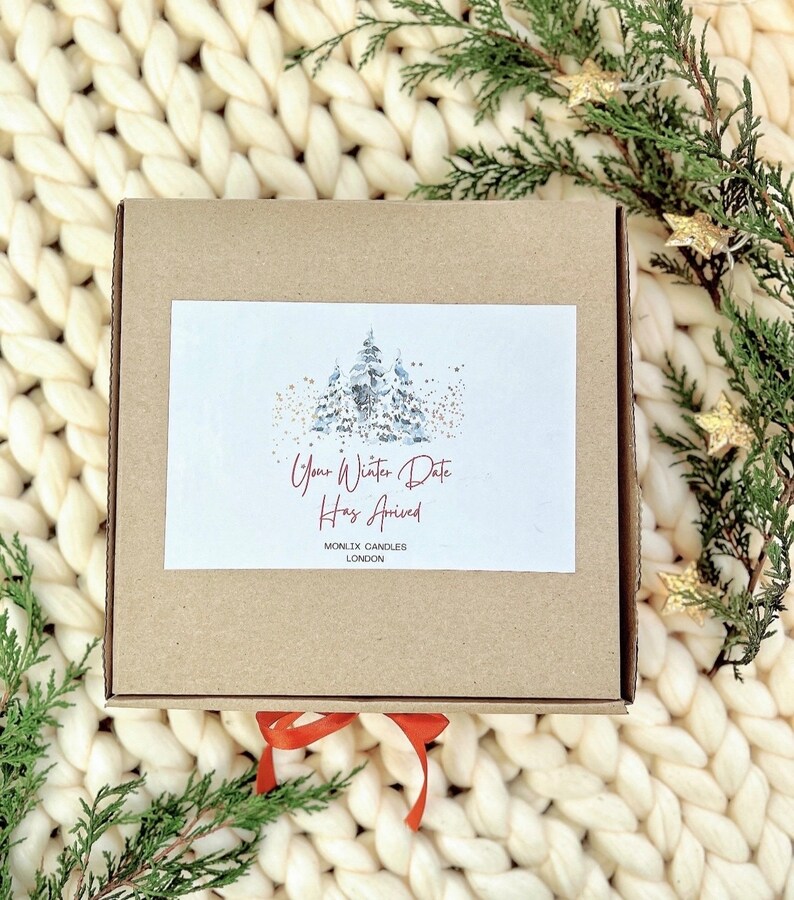 May include: A brown cardboard box with a white label that says "Your Winter Date Has Arrived" and "MONIX CANDLES LONDON". The box is sitting on a white knitted blanket with green pine branches and gold stars.