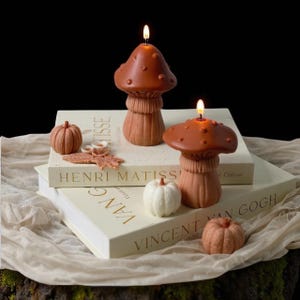 May include: Two lit mushroom-shaped candles in shades of brown sit atop books with the titles "Henri Matisse" and "Vincent Van Gogh." Small pumpkin and leaf-shaped decorations are scattered around the candles. The background is dark.