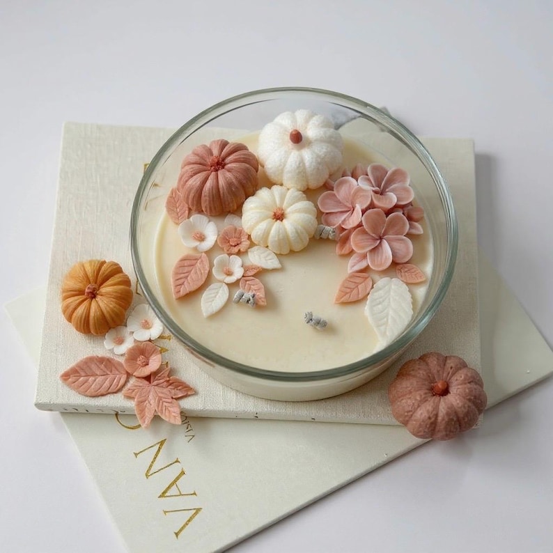 May include: A decorative candle in a clear glass bowl, adorned with miniature pumpkins, flowers, and leaves in shades of orange, white, and pink. The candle sits on a stack of books with gold lettering, creating a fall-themed aesthetic.