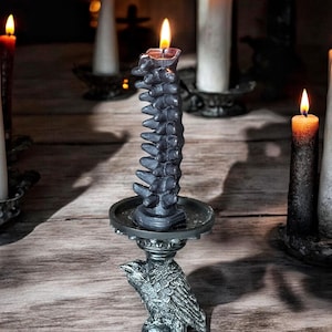 Pode incluir: Uma vela preta em forma de espinha dorsal est&aacute; sobre um casti&ccedil;al em forma de corvo. A vela est&aacute; acesa e o casti&ccedil;al &eacute; de metal.