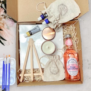 May include: A gift box filled with items for a paint and sip party. The box includes a small canvas, easel, paint tubes, a candle, a small bag of rose scented candles, a bottle of rose lemonade, and a small bag of rose petals.