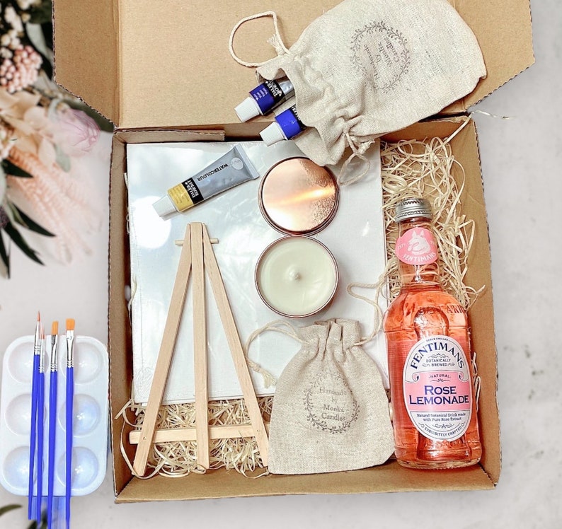 May include: A gift box filled with items for a paint and sip party. The box includes a small canvas, easel, paint tubes, a candle, a small bag of rose scented candles, a bottle of rose lemonade, and a small bag of rose petals.
