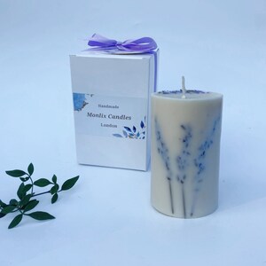 May include: A white candle with dried lavender flowers embedded in the wax. The candle is in a white box with a label that says "Handmade Monlix Candles London".