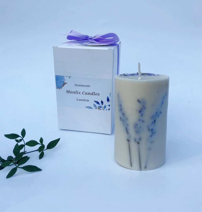 May include: A white candle with dried lavender flowers embedded in the wax. The candle is in a white box with a label that says "Handmade Monlix Candles London".