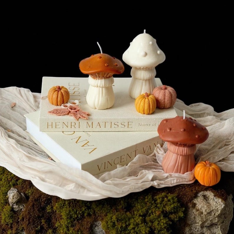 May include: A collection of mushroom-shaped candles in shades of brown, white, and peach, alongside miniature orange pumpkins. The candles are arranged on books with the text "HENRI MATISSE" and "VINCENT VAN".