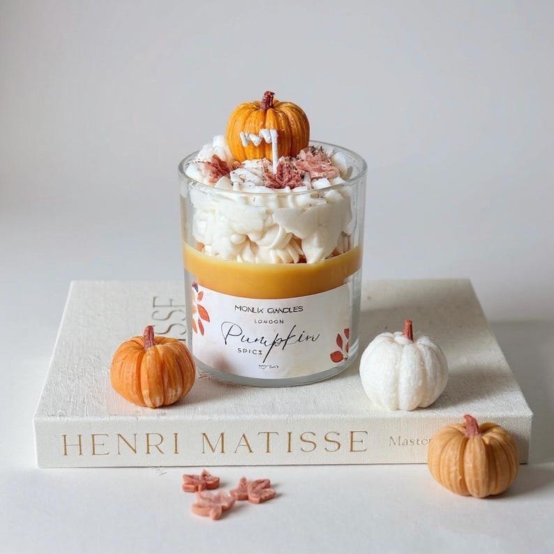 Puede incluir: Una vela de especias de calabaza en un tarro de cristal transparente, cubierta con glaseado blanco y una peque&ntilde;a calabaza naranja. La vela se encuentra sobre un libro blanco con las palabras "HENRI MATISSE" y varias calabazas decorativas peque&ntilde;as.