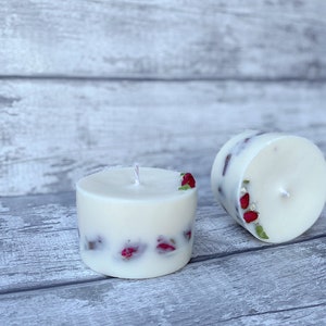 May include: Two white pillar candles with dried red flowers embedded in the wax. The candles are on a light gray wooden surface.